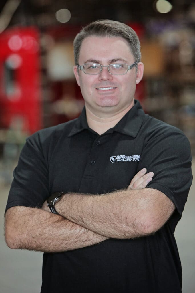 A man with glasses and a black polo shirt, with his arms crossed, smiling at the camera.