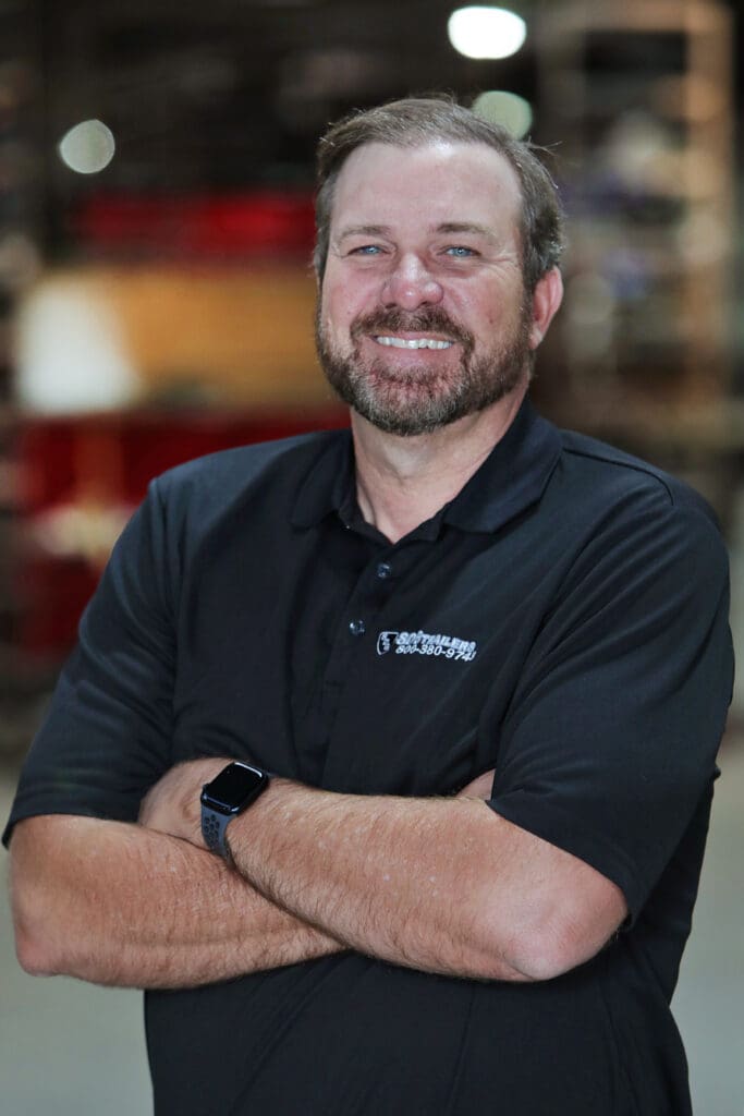 A man with a beard and short hair, wearing a black polo shirt with a logo, stands with his arms crossed and smiles at the camera.