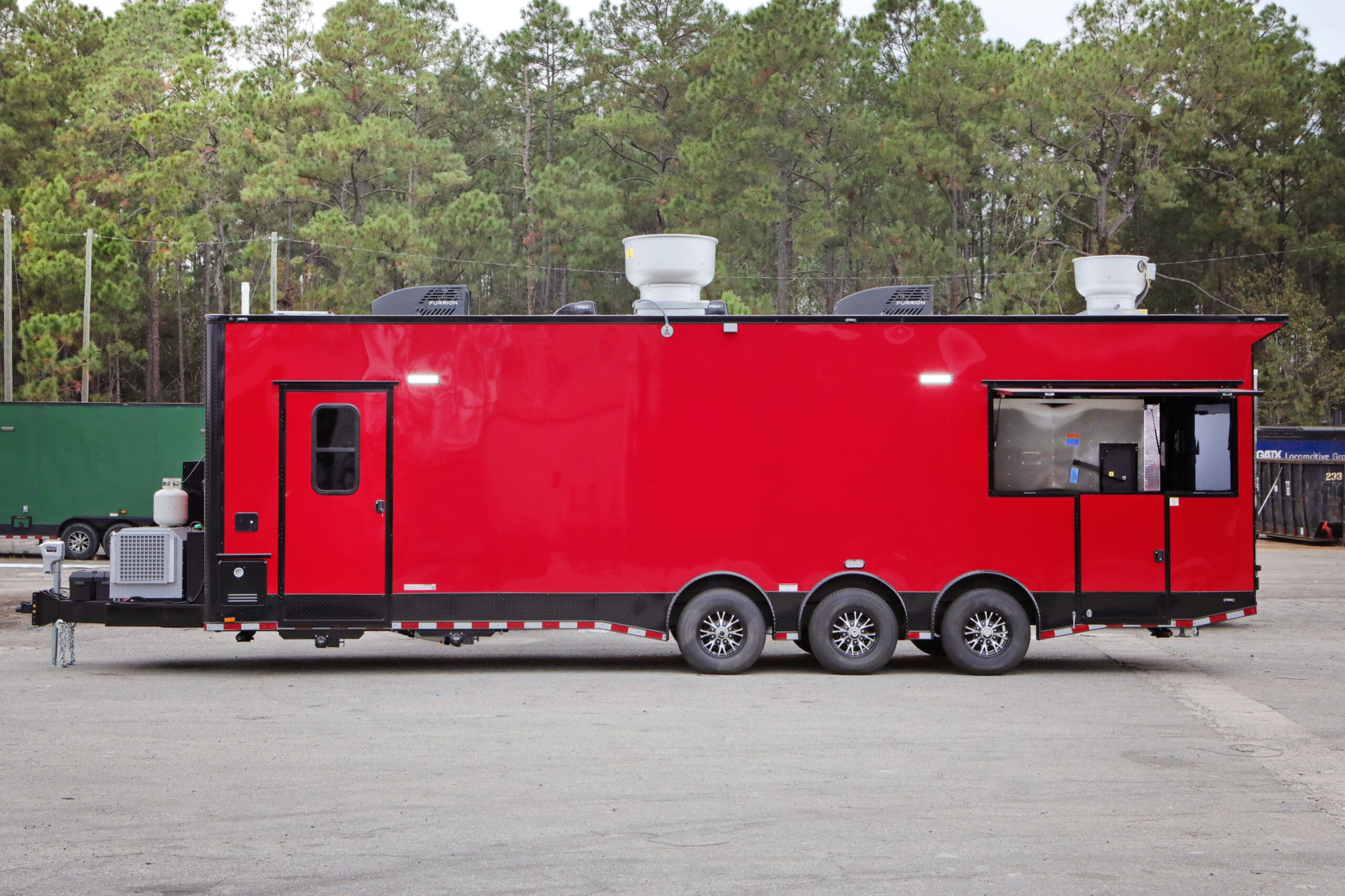 A red food or BBQ trailer with multiple axles and exhaust vents on the roof, parked outdoors with trees in the background.