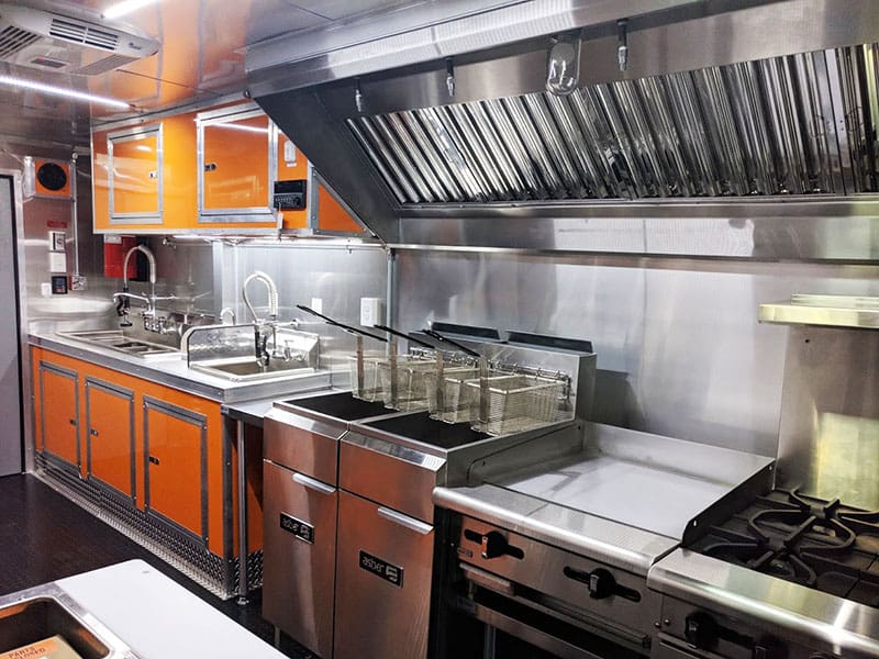 Interior view of a commercial kitchen inside a food truck or concession trailer, featuring stainless steel countertops, deep fryers, a stove with an oven, a range hood, and orange cabinetry along one side